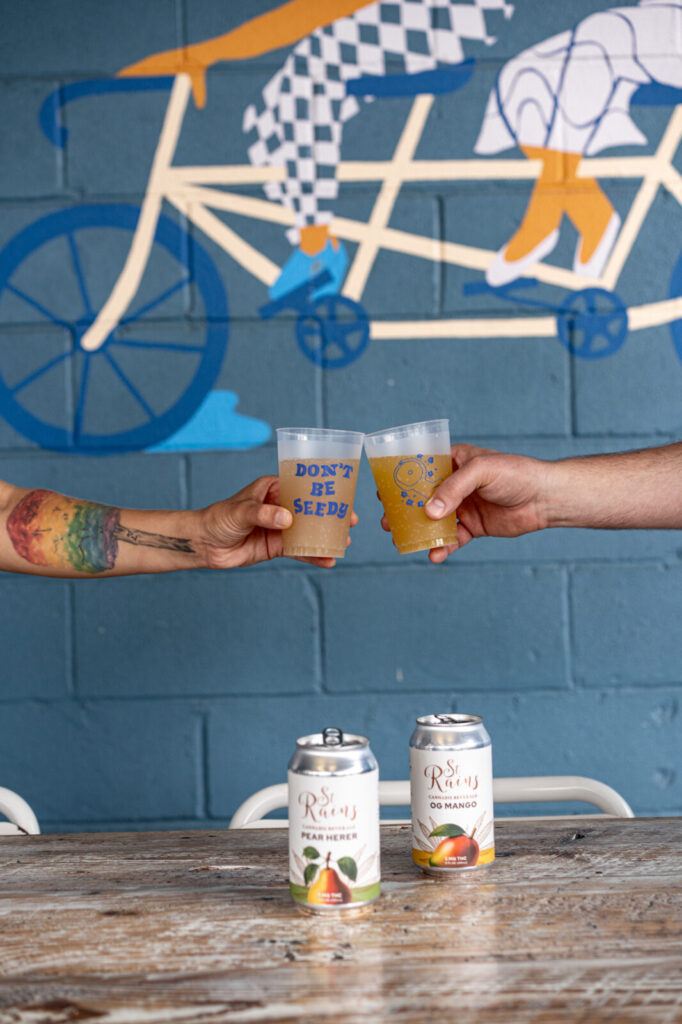 Two cups of St. Rains toasted in front of the blue bike mural at Flour Moon Bagels