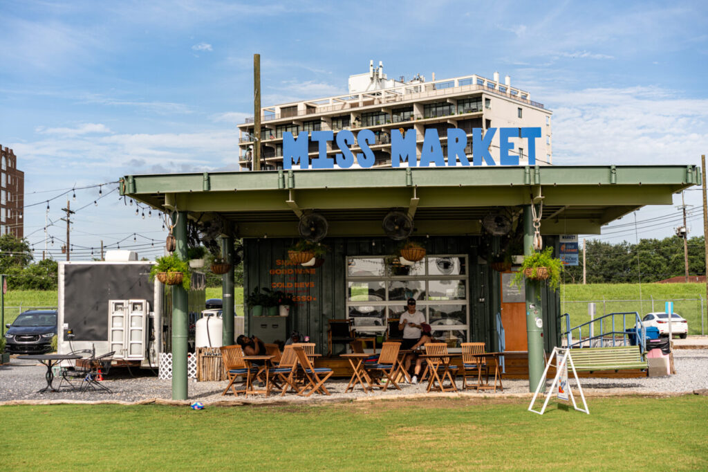 Exterior of Miss Market at the Batture with blue lettering, an open-air pavilion, and picnic seating on the lawn