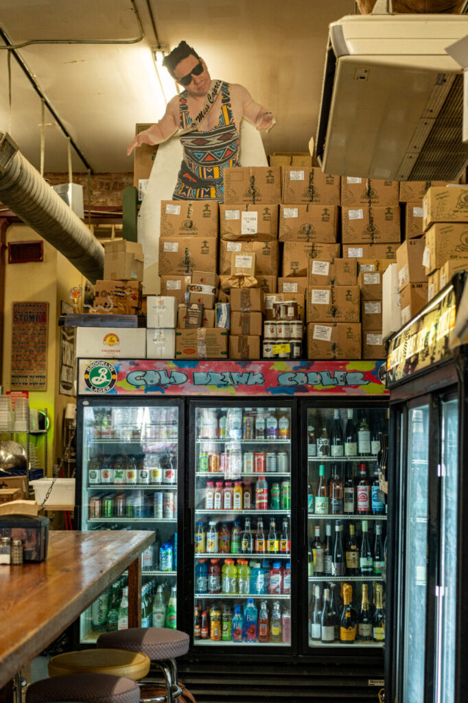 Cold drink coolers at Stein's Deli stocked with bottles and cans, stacked boxes above