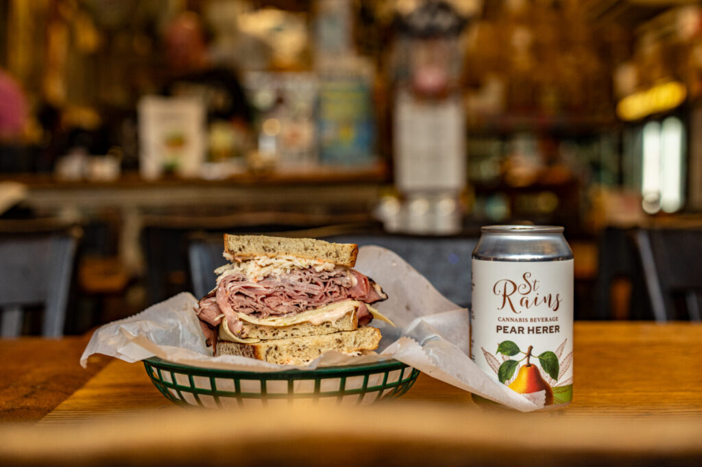 Stein's Deli sandwich piled with meat and slaw in a basket, next to a can of St. Rains Pear Herer